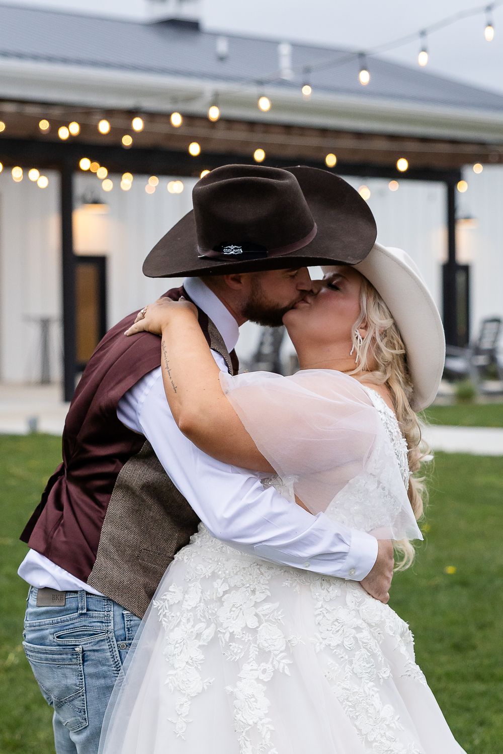 Bride and groom sharing a kiss in cowboy hats at an outdoor wedding venue with string lights.