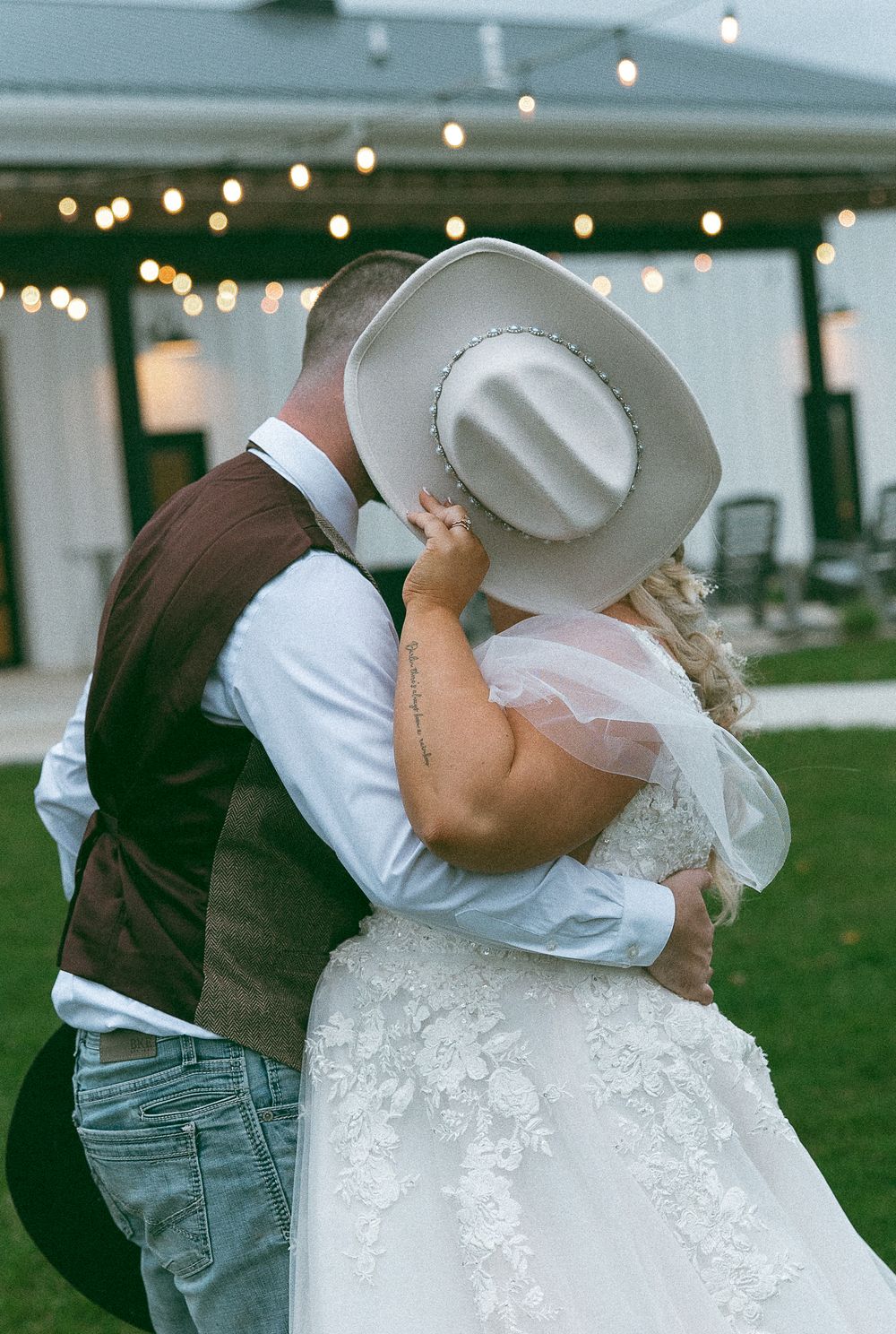 Bride and groom share a romantic kiss hidden beneath a white cowboy hat at an outdoor wedding reception.