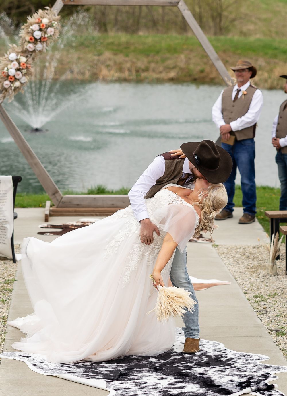 Bride and groom share a romantic dip kiss at outdoor country wedding ceremony by a pond with hexagon arch.