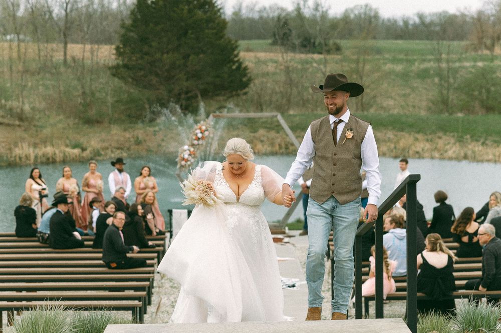 Bride and groom walk down outdoor wedding aisle holding hands, with guests seated and a pond in the background.