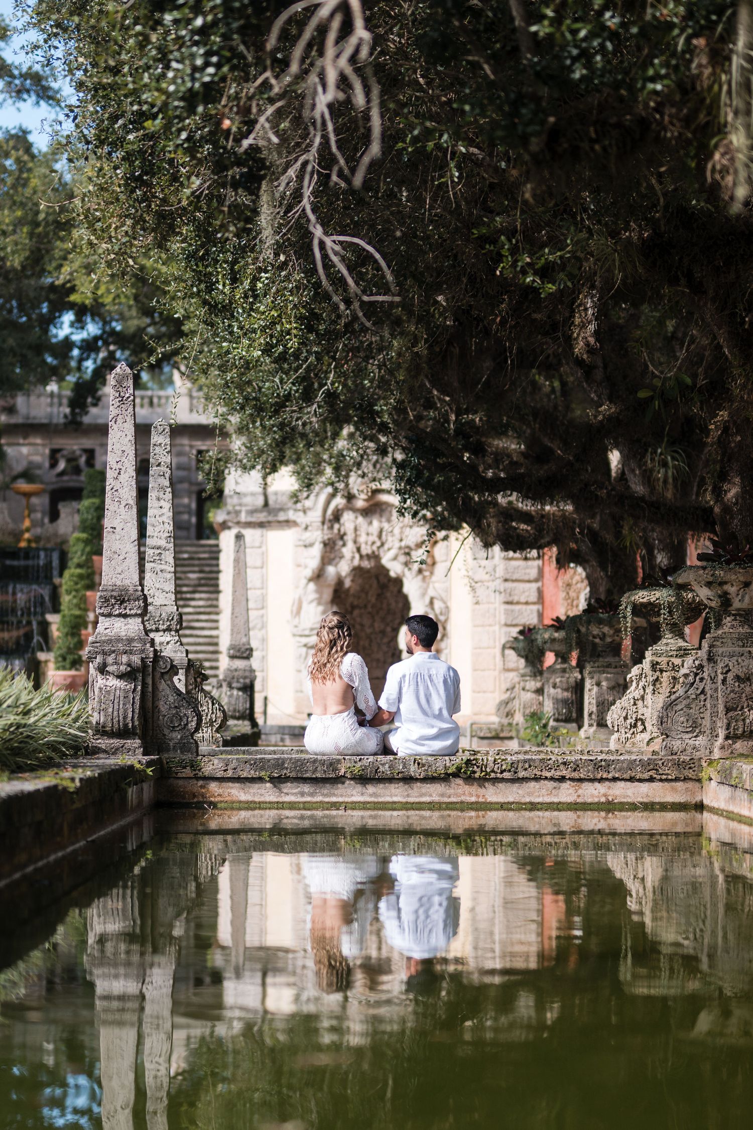 Bride and groom walk through Vizcaya Gardens beside a lily pond, surrounded by tropical greenery and historic stone columns.