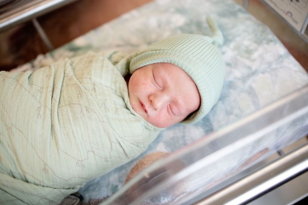 Sleeping newborn baby swaddled in green blanket wearing knit hat in hospital bassinet.