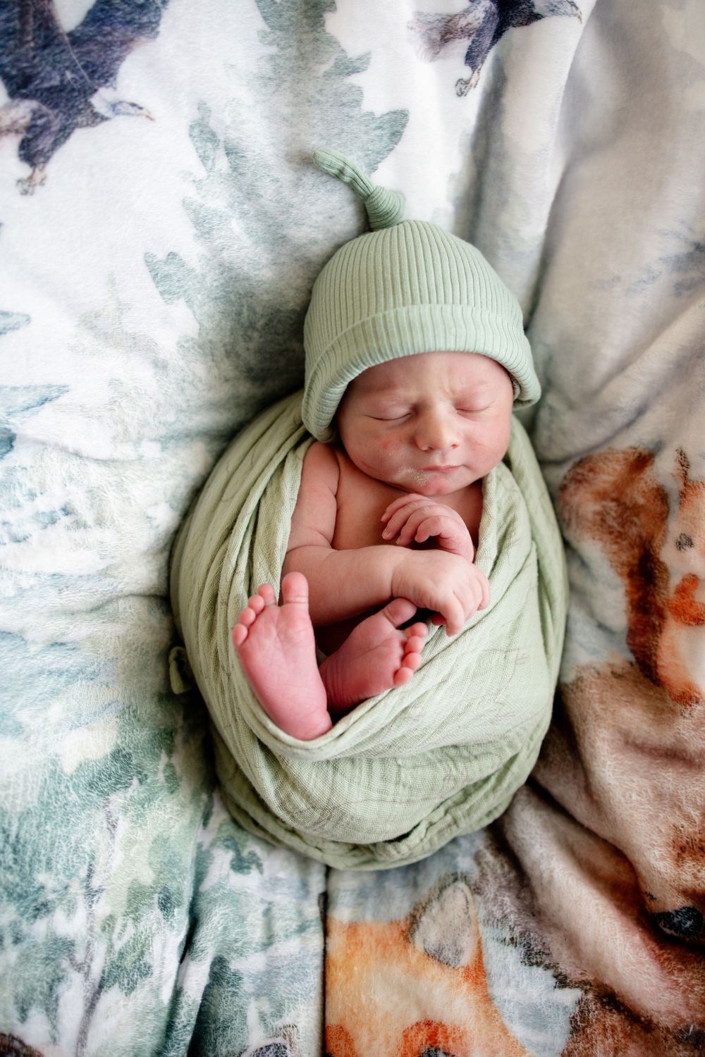 Newborn baby swaddled in green wrap wearing knit hat, sleeping peacefully on floral blanket.