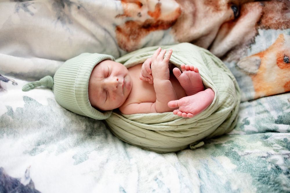 Sleeping newborn baby wrapped in soft green knit swaddle and matching hat posed on woodland animal print blanket.