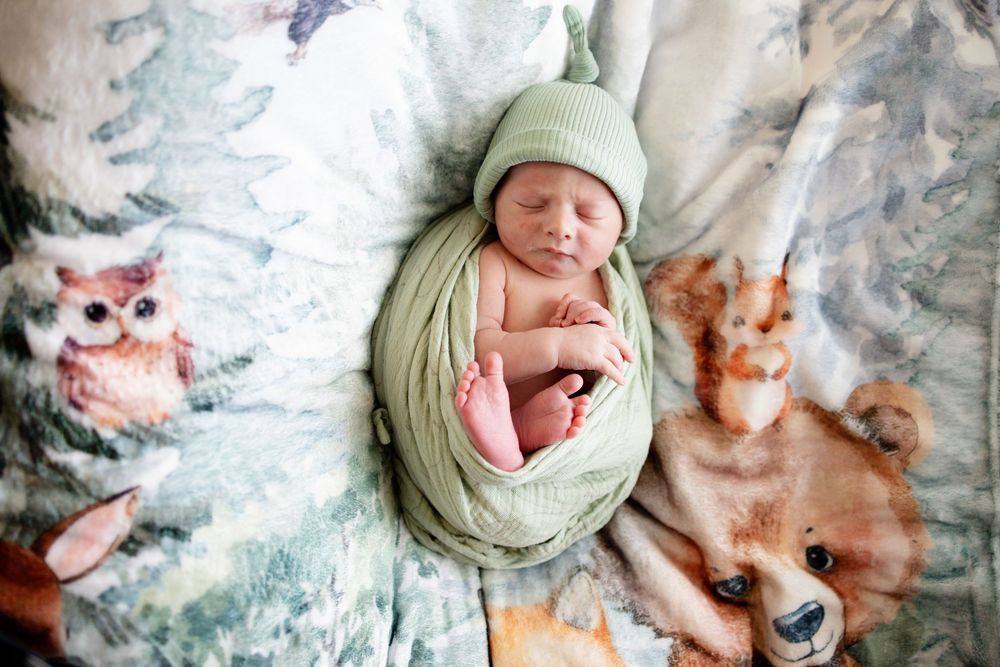 Sleeping newborn baby wrapped in green swaddle with knit hat on woodland animal blanket.