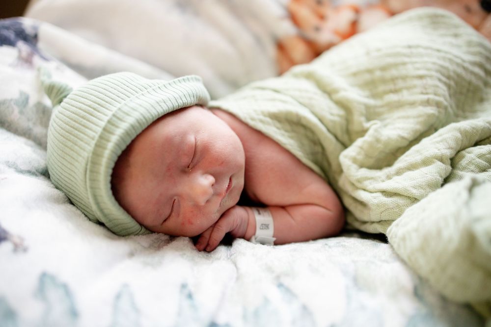 Peaceful newborn baby sleeping swaddled in green knit blanket and matching hat on patterned bedding.