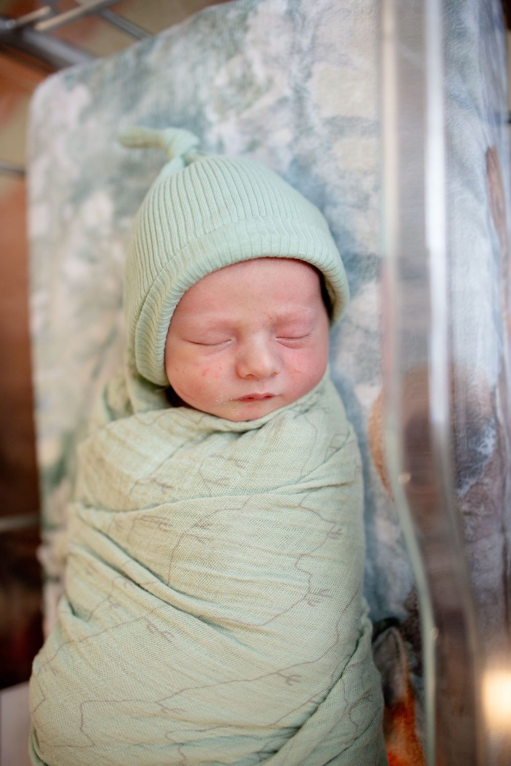 Swaddled newborn baby sleeping peacefully in hospital bassinet wearing sage green knit hat.