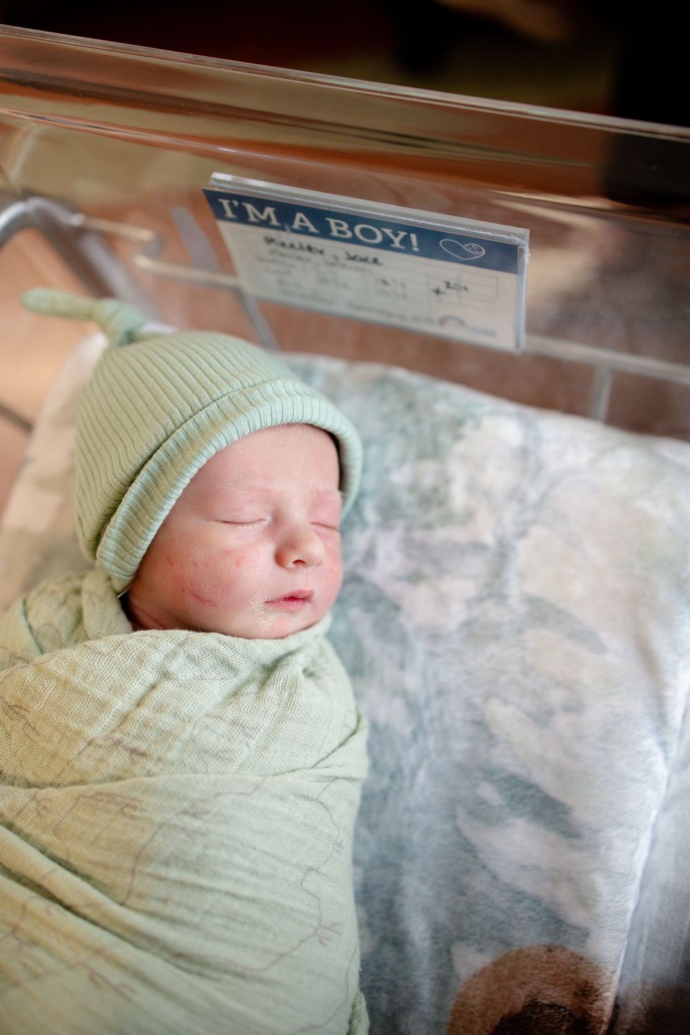 Newborn baby boy swaddled in green blanket wearing knit hat sleeping in hospital bassinet with I'm a Boy sign.