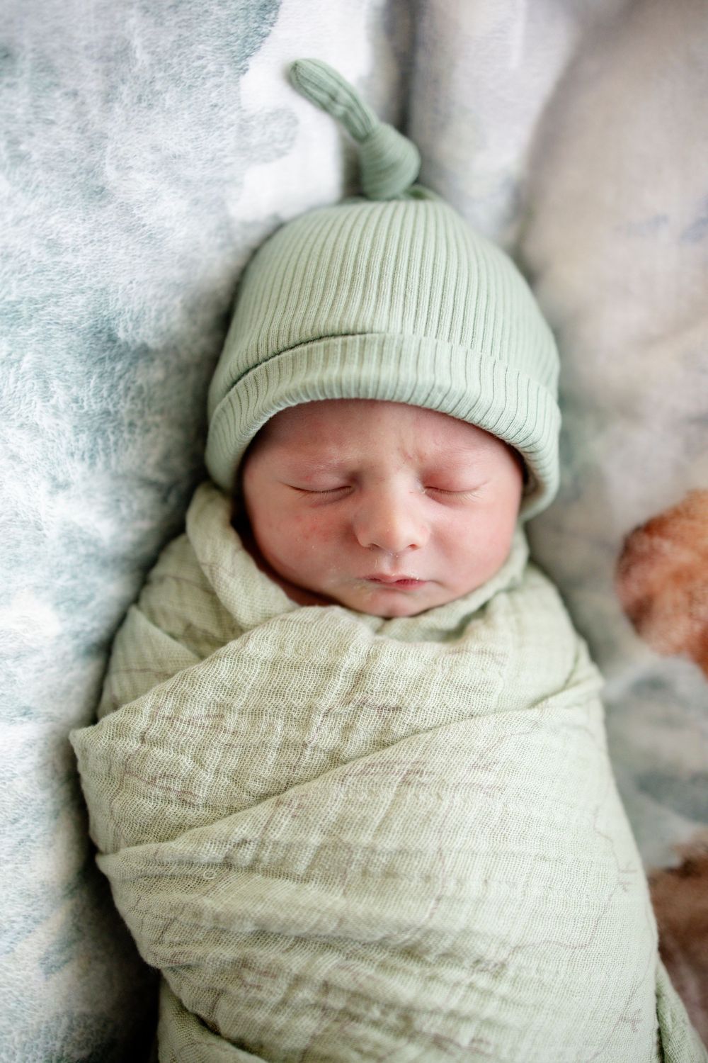 Sleeping newborn baby tightly swaddled in light green muslin wrap wearing matching knotted beanie hat.