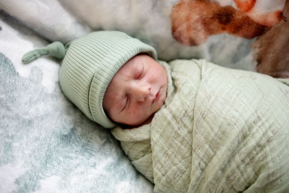 Peaceful newborn baby wrapped in green muslin swaddle wearing sage knit hat sleeping.