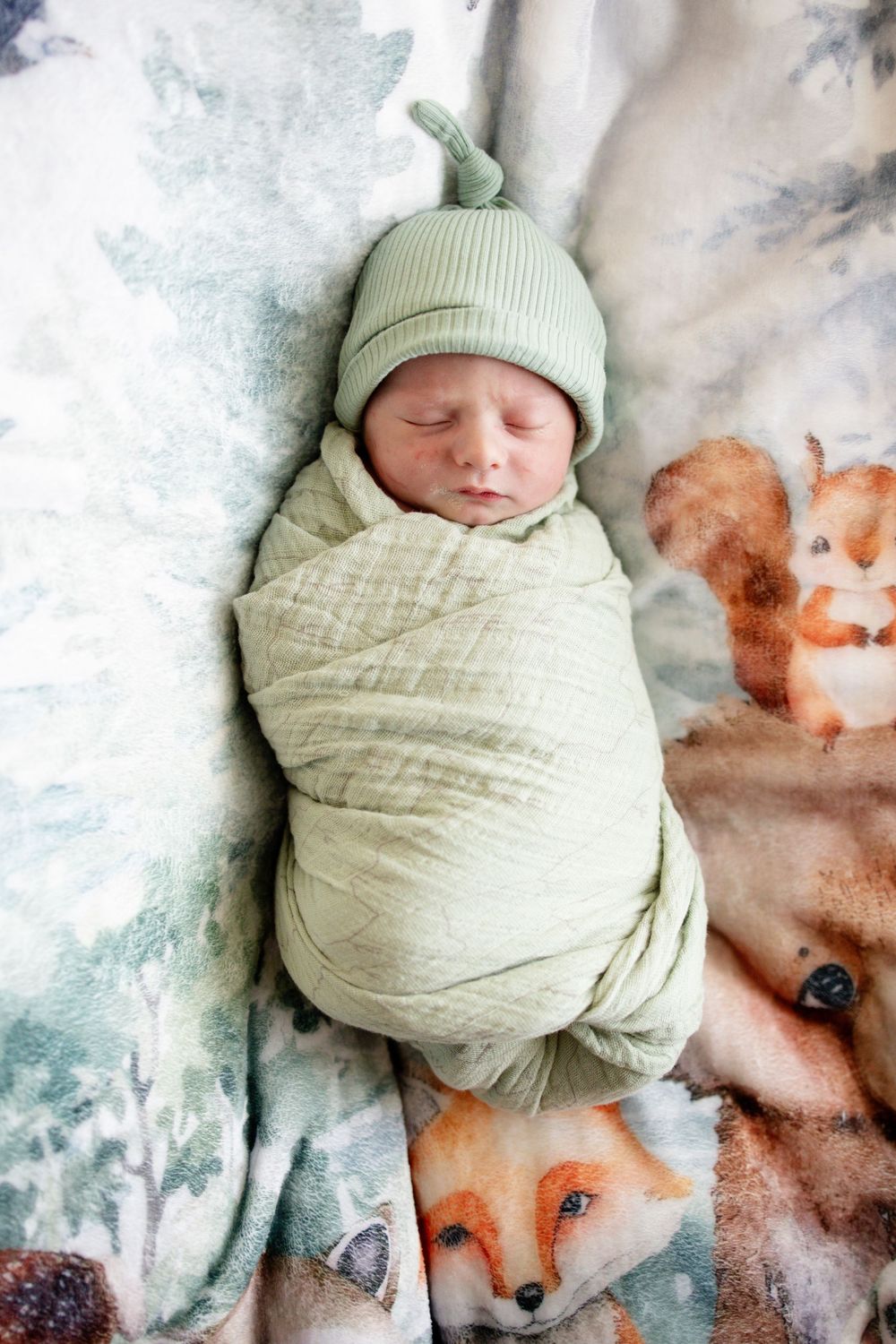 Newborn baby swaddled in sage green wrap sleeping on woodland animals blanket with fox and squirrel.