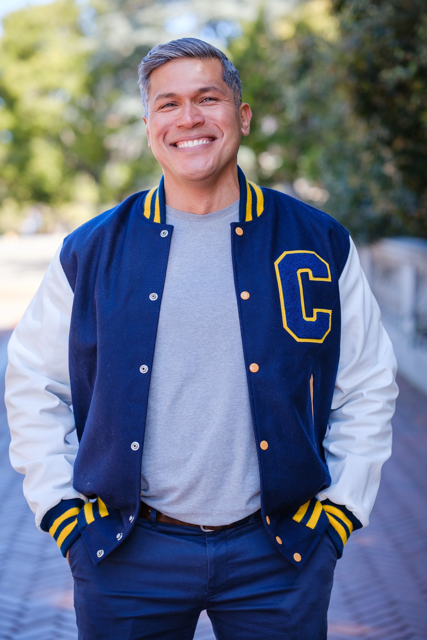 Smiling middle-aged man wearing a blue varsity jacket with white sleeves and yellow C letter, standing outdoors.