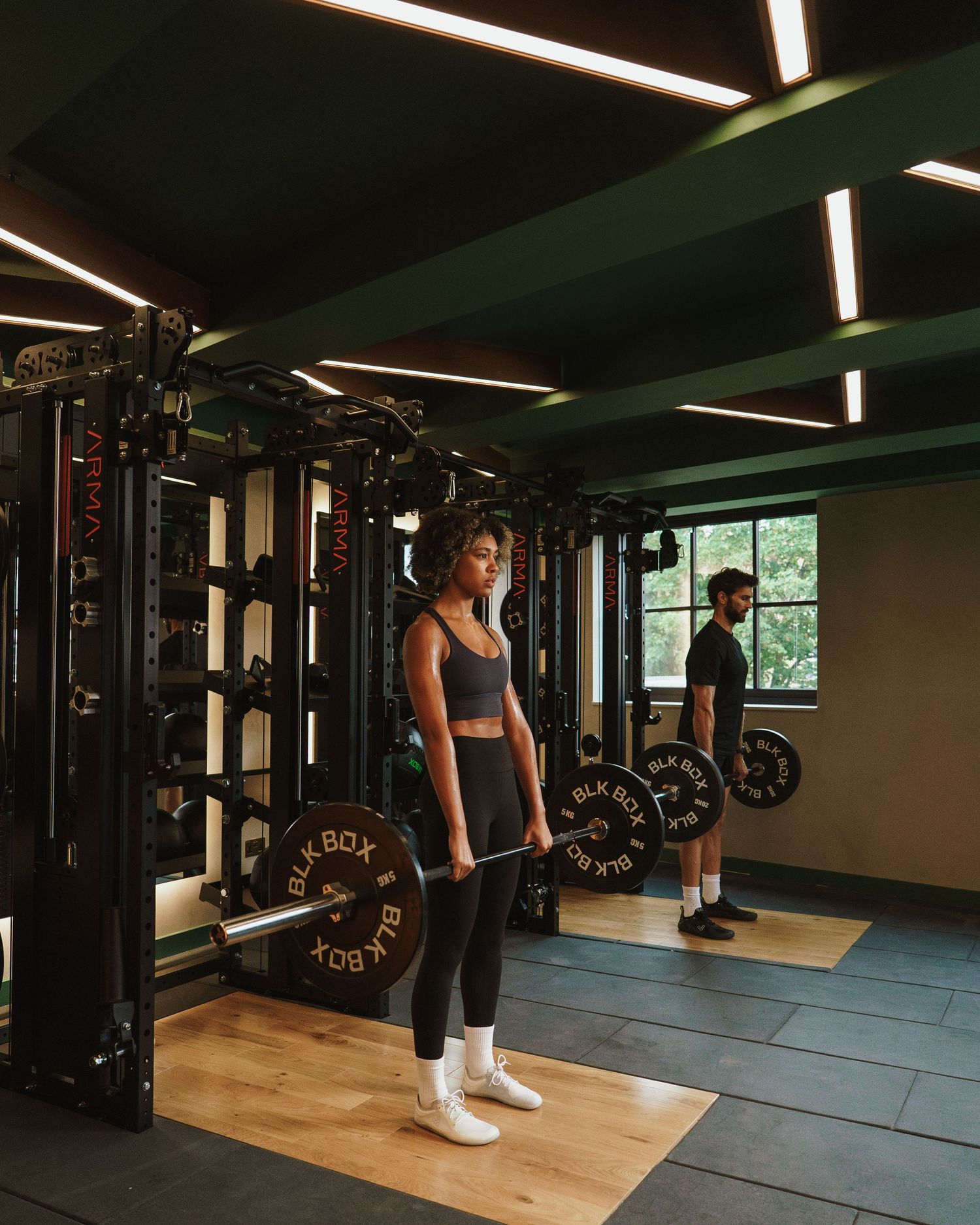 Two people lifting barbells in a modern gym with black equipment, green ceiling beams, and wooden flooring.