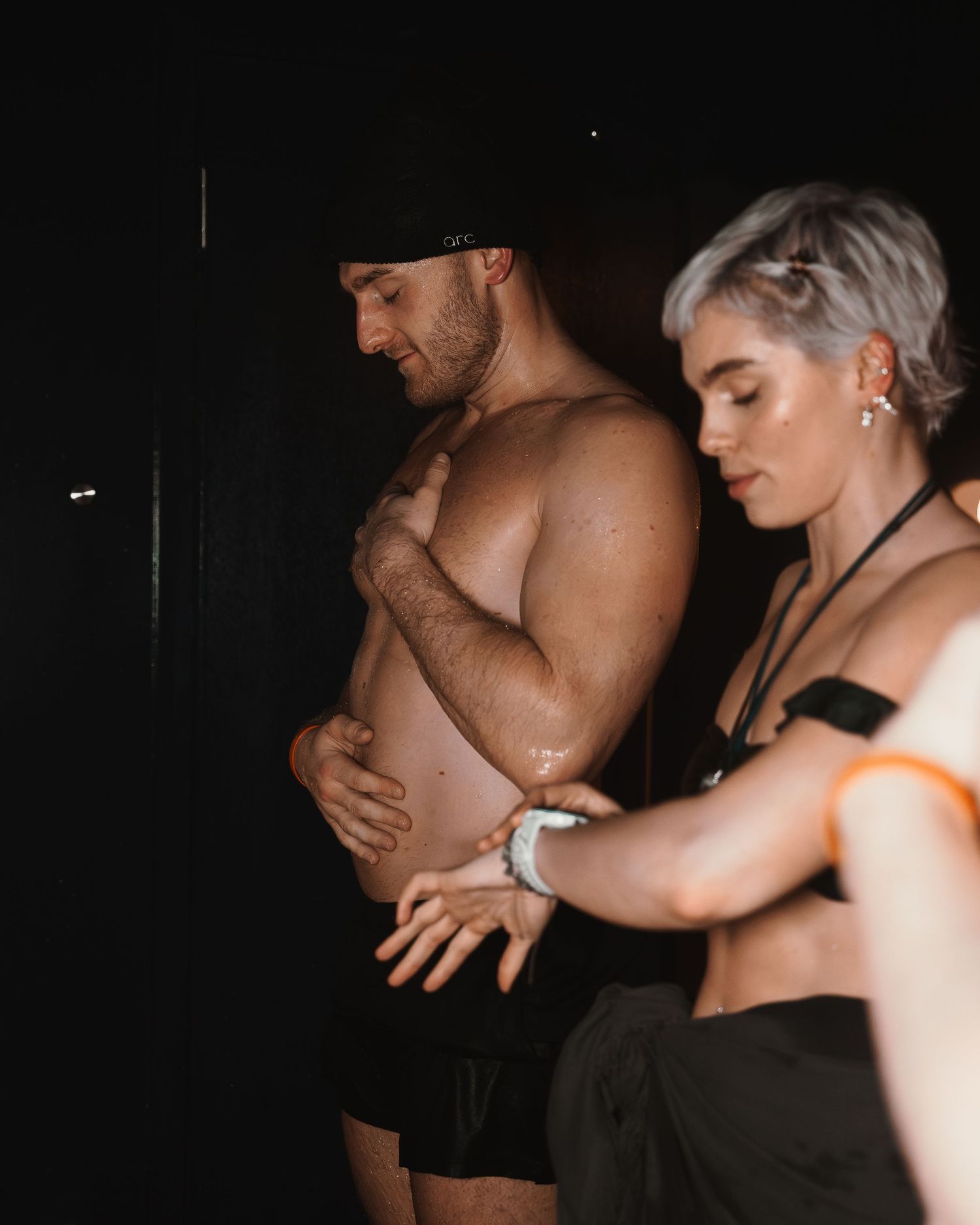 A shirtless muscular man and a woman with silver hair in a dark setting at an event.
