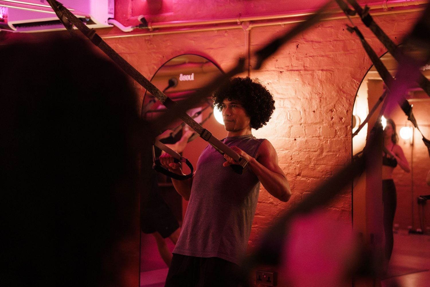 Woman with afro hair exercises using TRX suspension straps in a pink-lit gym with brick walls.