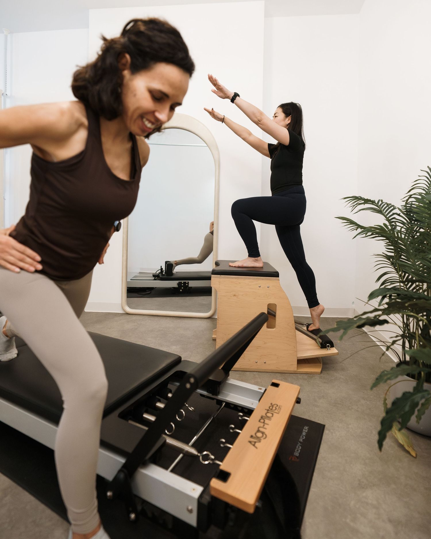 Two women practicing Pilates in a modern studio with reformer equipment, ladder barrel, and mirror.