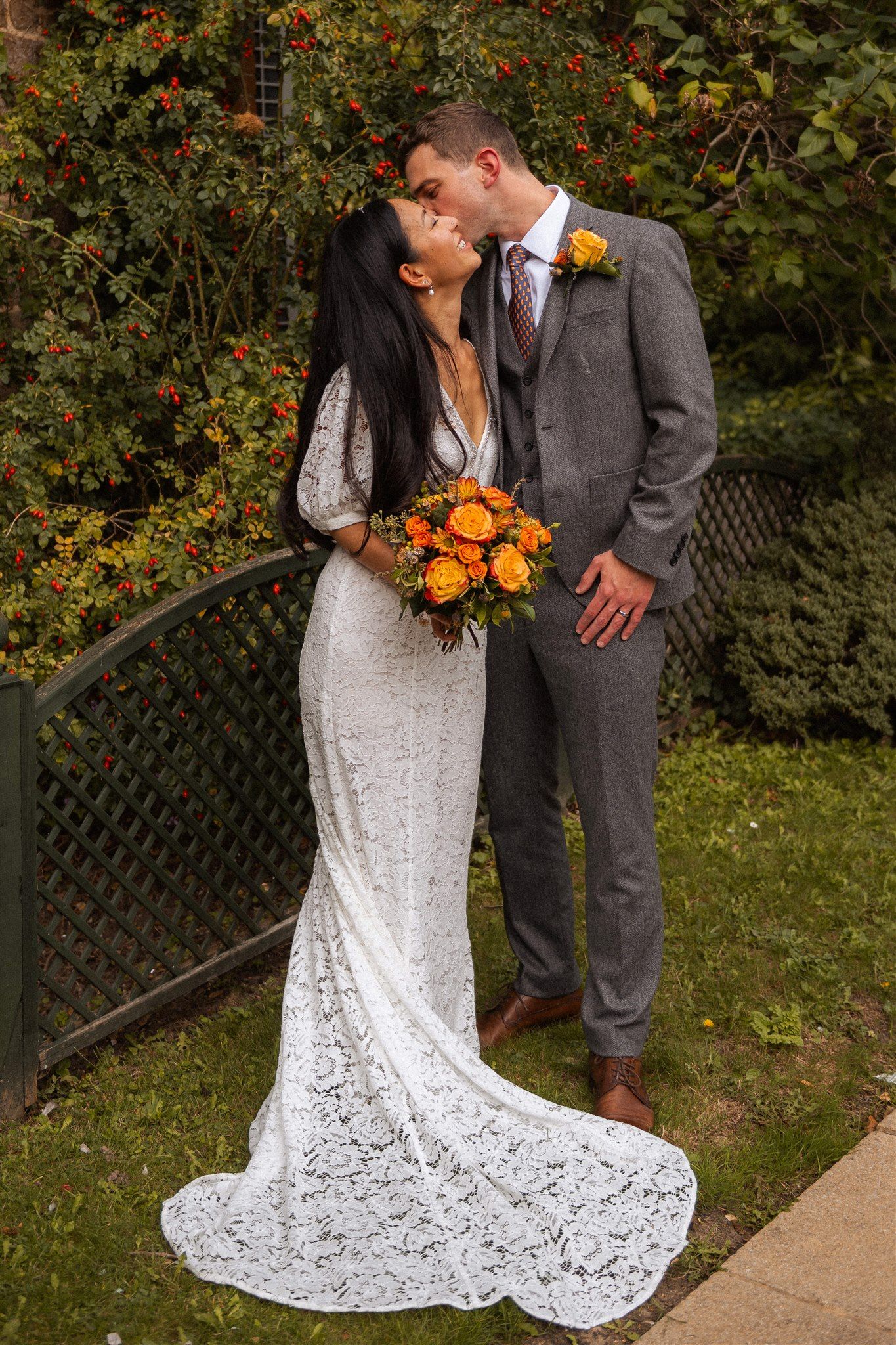 Bride in lace gown and groom in grey suit share a kiss outdoors, holding orange rose bouquet with autumn foliage backdrop.