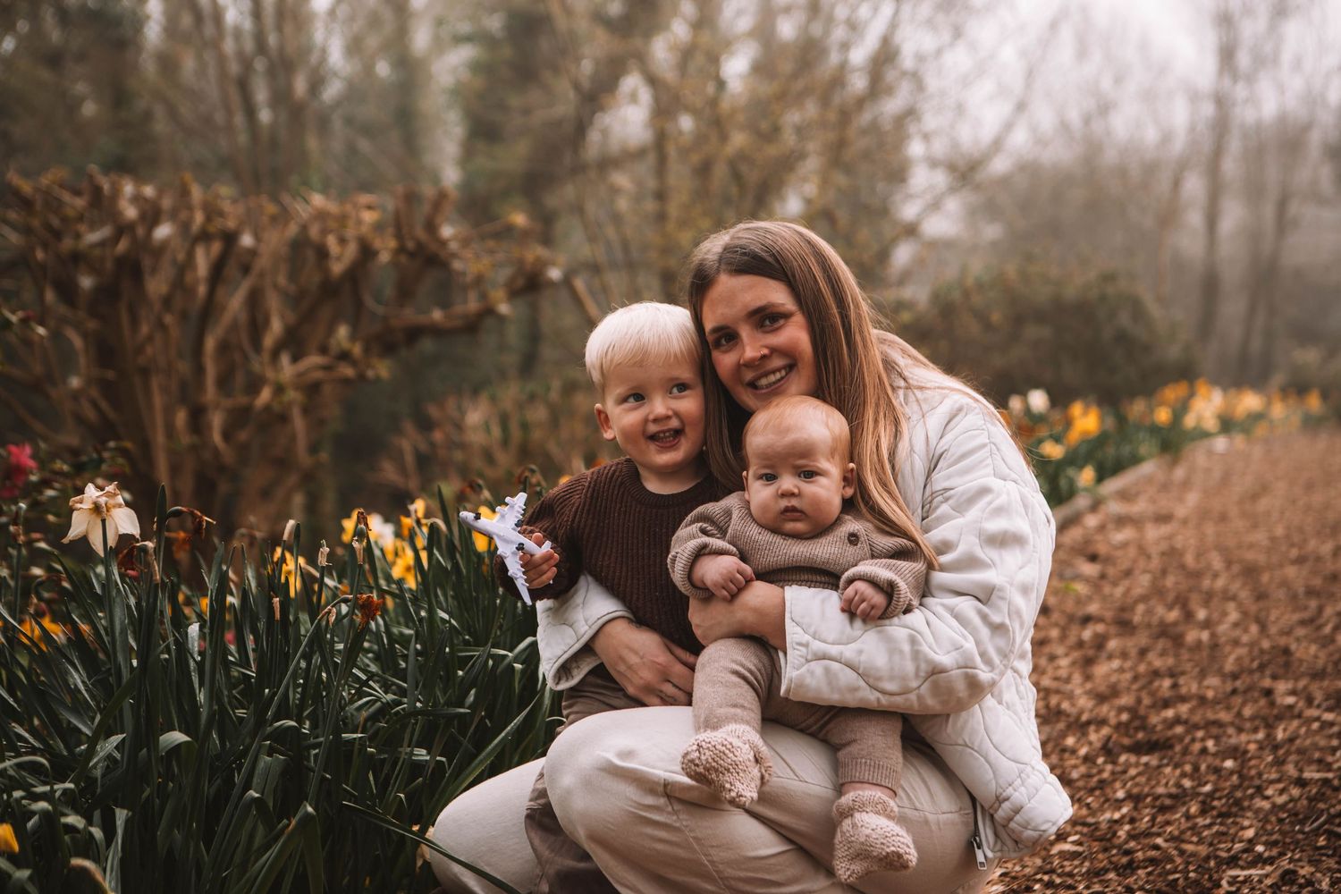 A smiling mother sits outdoors with two young children among spring daffodils in a garden path setting.