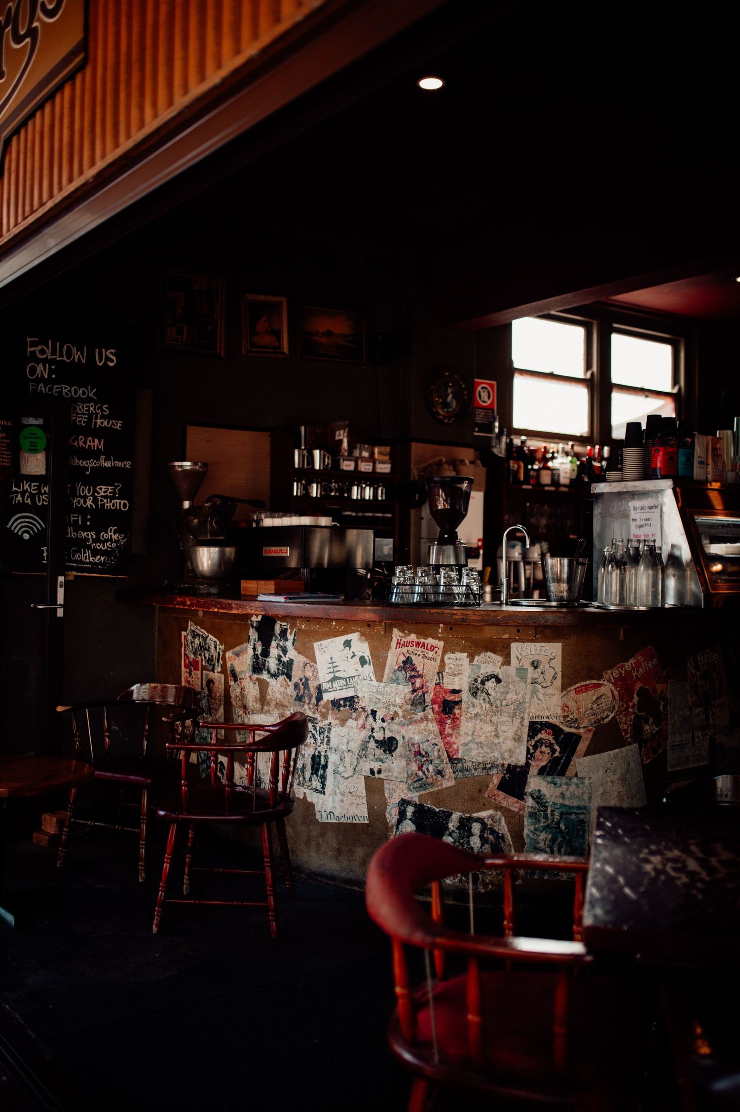 Dimly lit vintage cafe interior with rustic wooden chairs, a bar covered in posters, chalkboard signs, and warm ambient lighting.