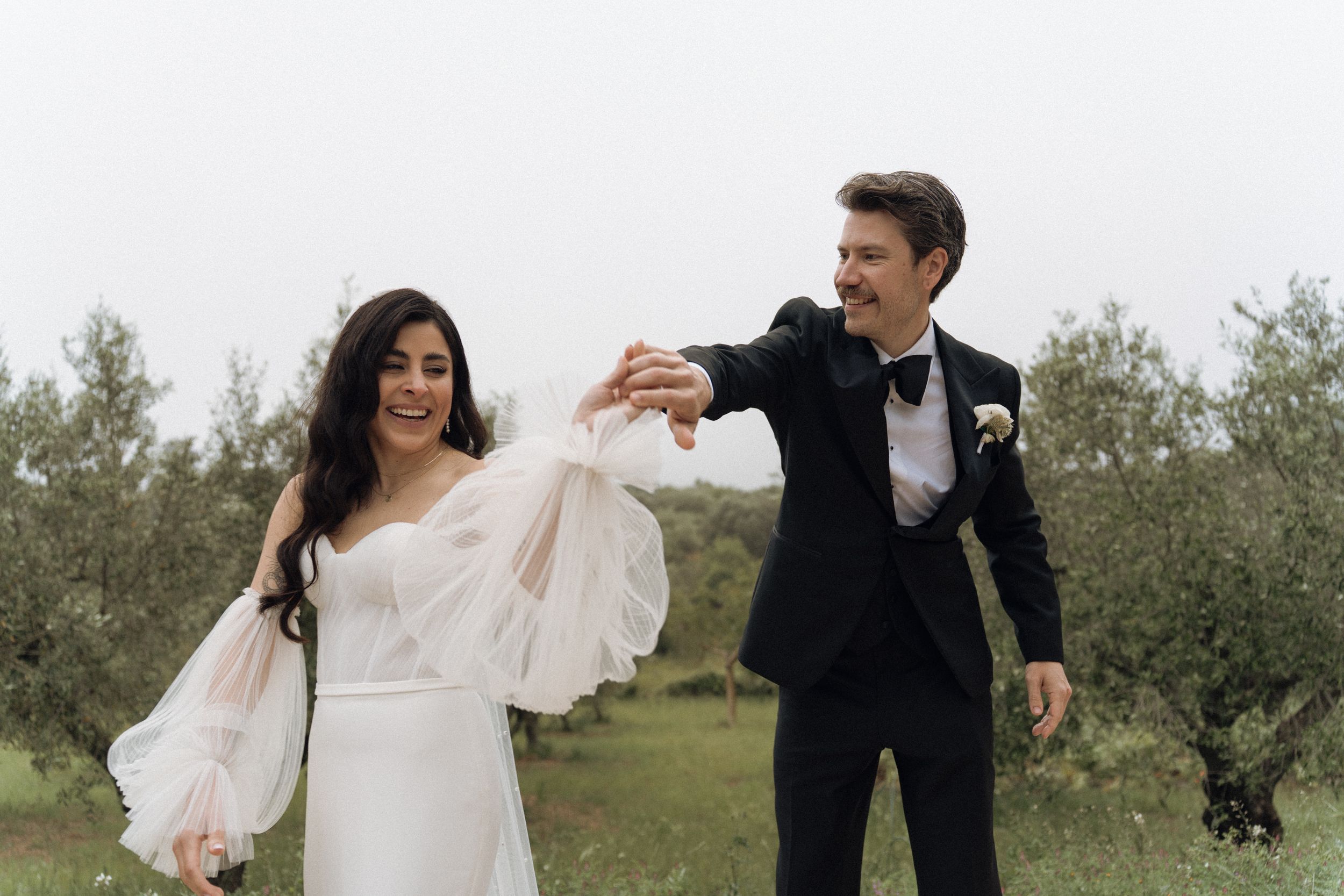 Bride in white gown and groom in black tuxedo holding hands and dancing outdoors among olive trees.