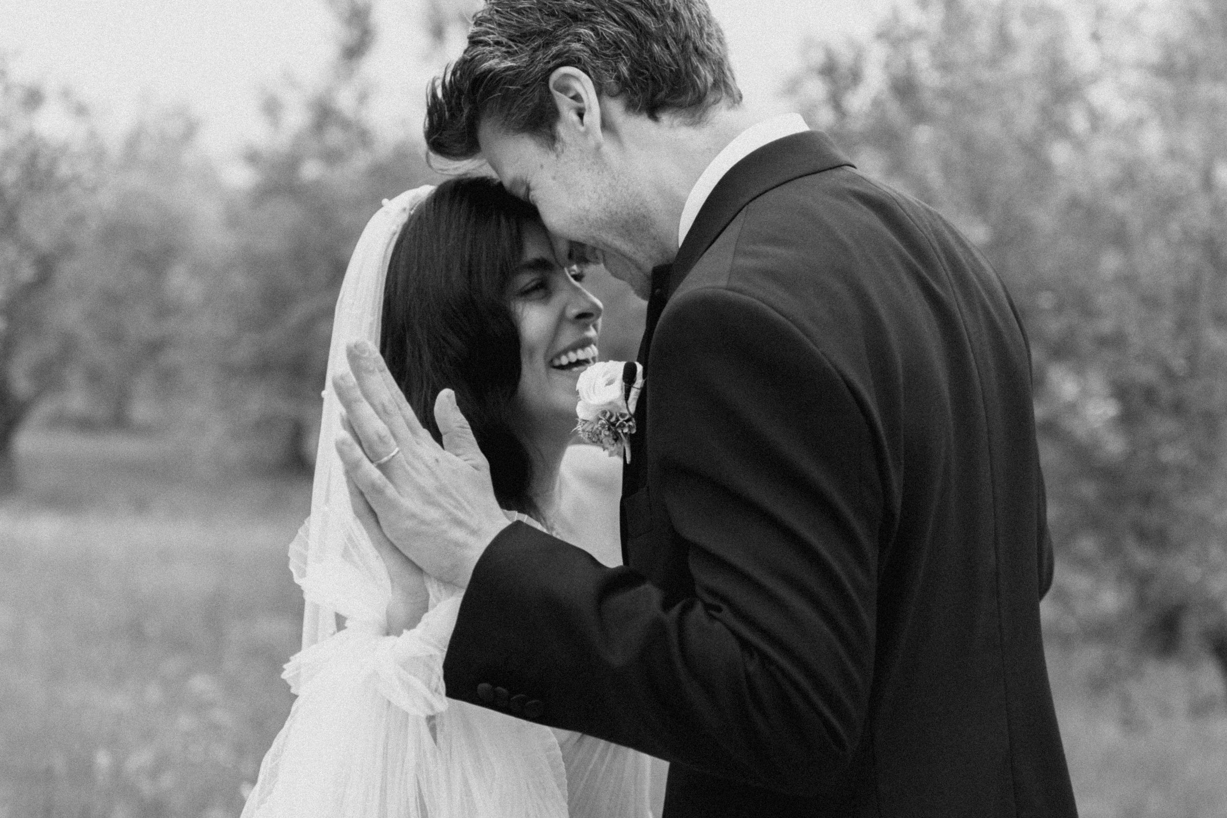 Black and white wedding photo of smiling bride in veil and groom in suit sharing a tender moment outdoors.