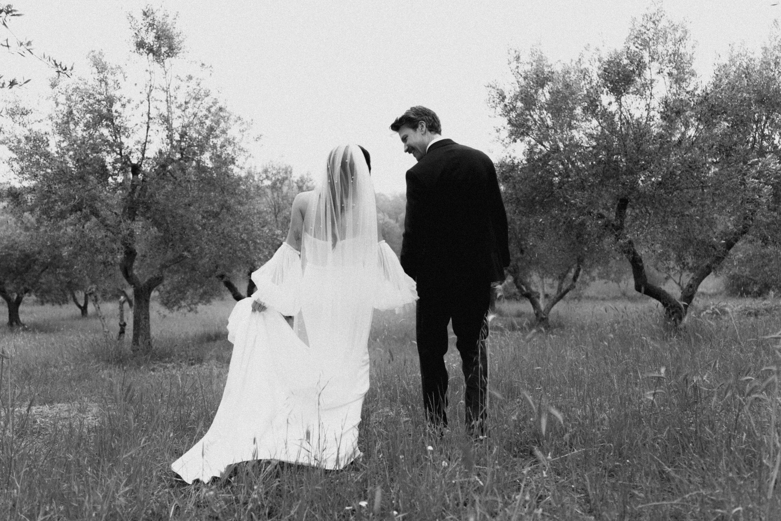 Black and white photo of a bride and groom walking hand in hand through an olive grove on their wedding day.