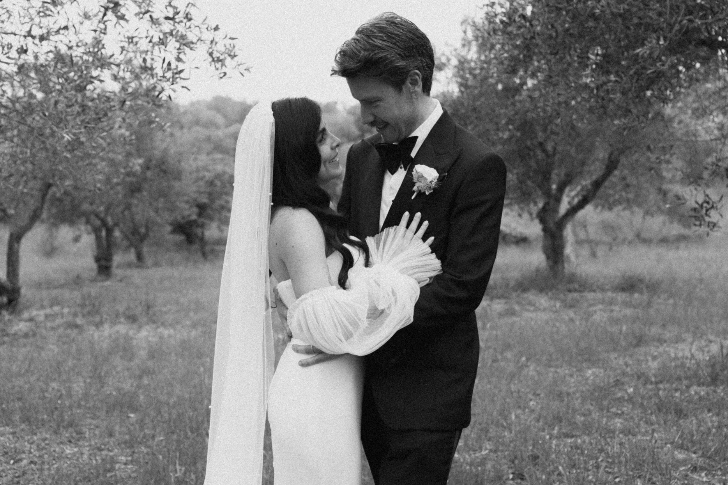 Black and white wedding photo of bride and groom embracing in an olive grove, bride holding white bouquet.