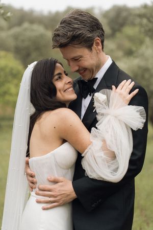 Bride and groom embrace outdoors, smiling at each other, wearing wedding dress with veil and black tuxedo.