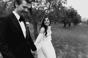 Black and white photo of a smiling bride and groom walking among olive trees on their wedding day.