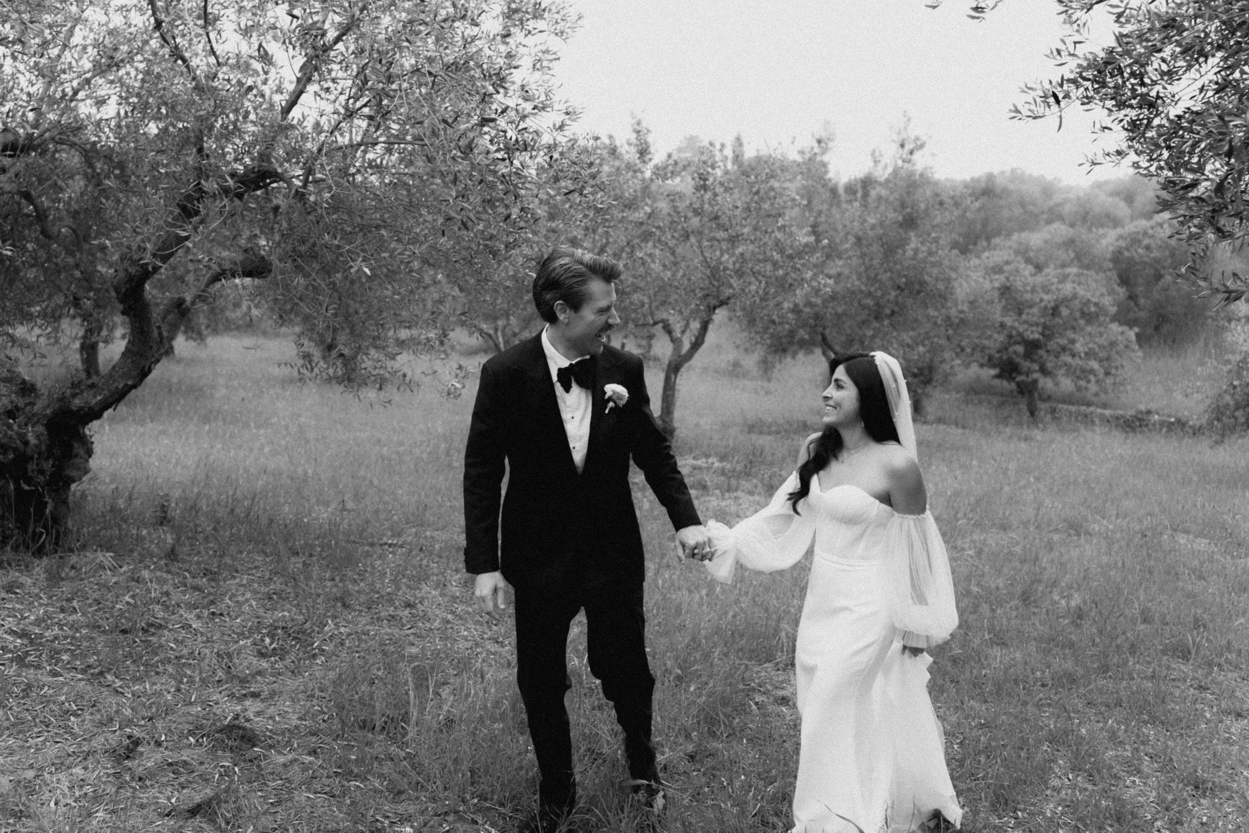 Black and white photo of a bride and groom holding hands walking through an olive grove on their wedding day.