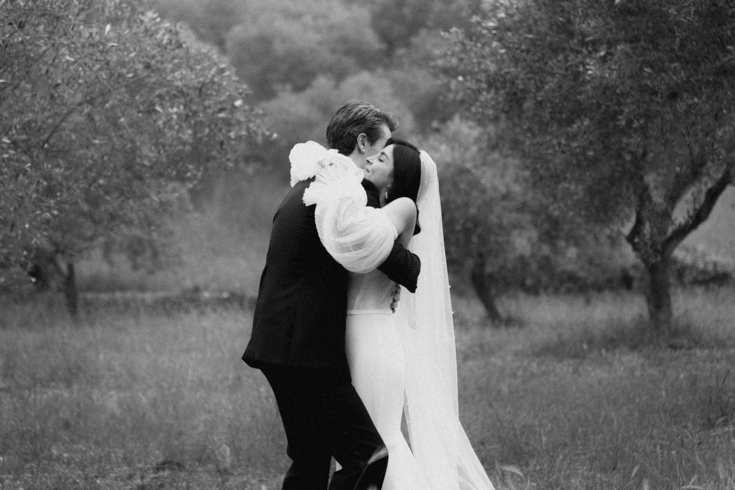 Black and white photo of a bride and groom sharing a romantic kiss in a lush outdoor garden setting.
