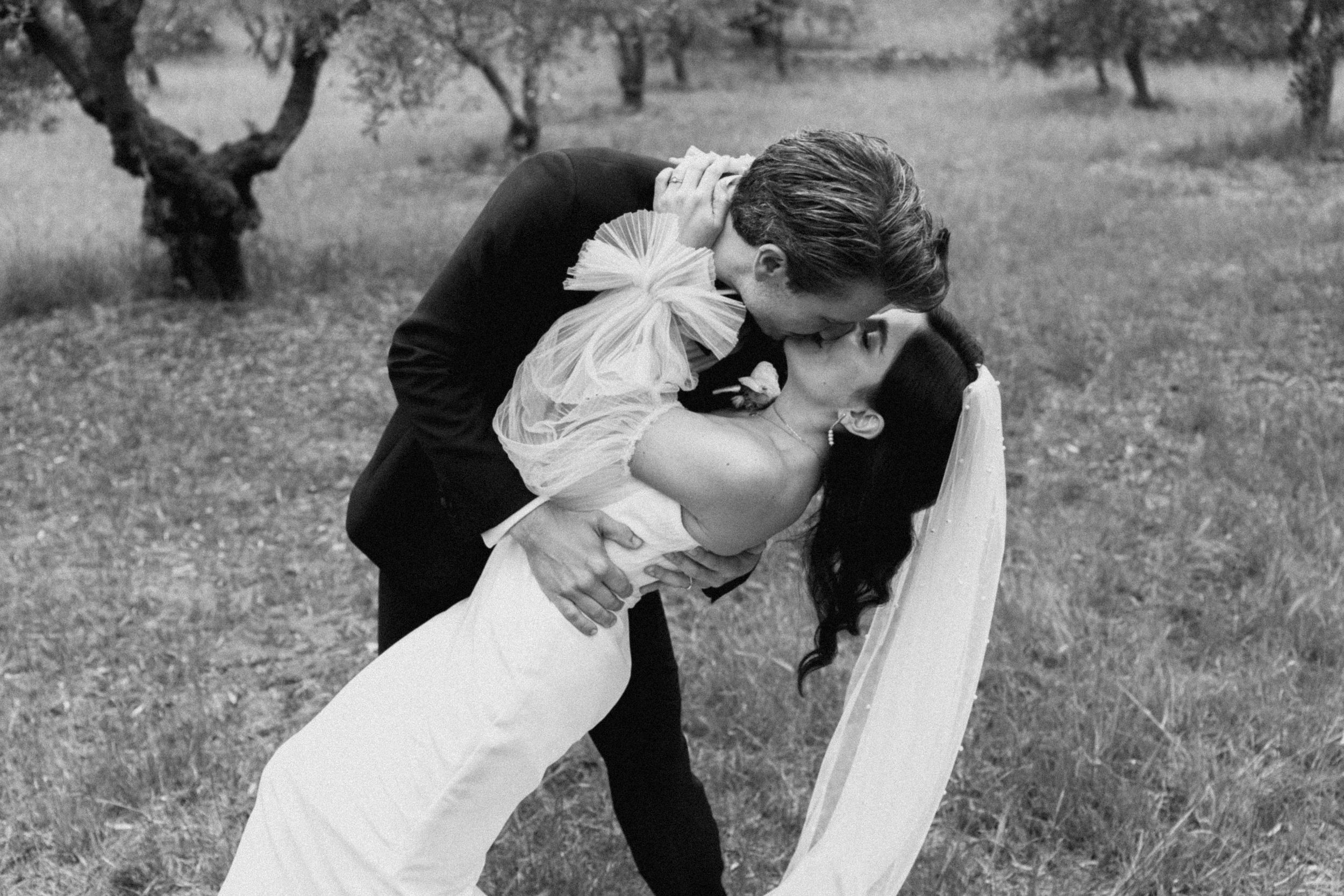 Black and white photo of a groom dipping and kissing his bride outdoors among olive trees on their wedding day.