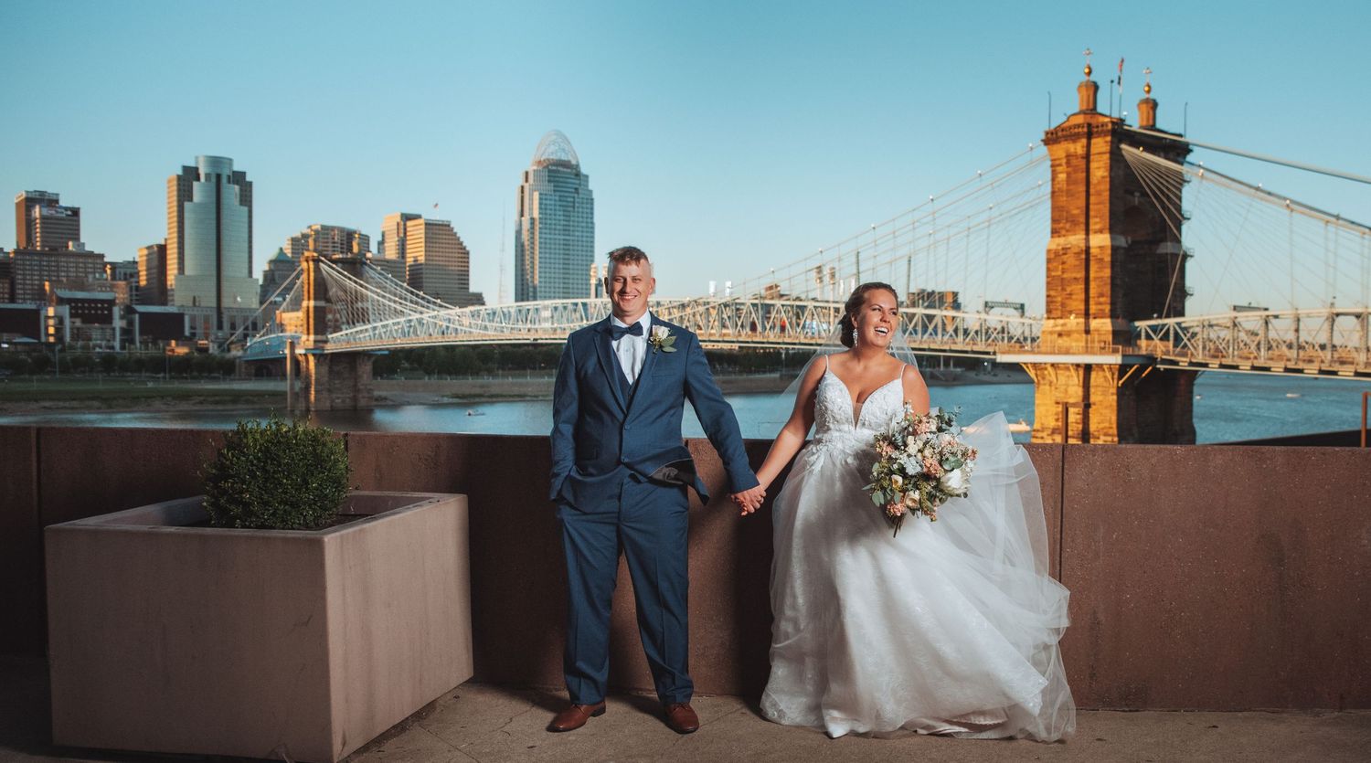 Bride and groom hold hands on a Cincinnati riverfront terrace with the Roebling Bridge and skyline backdrop.