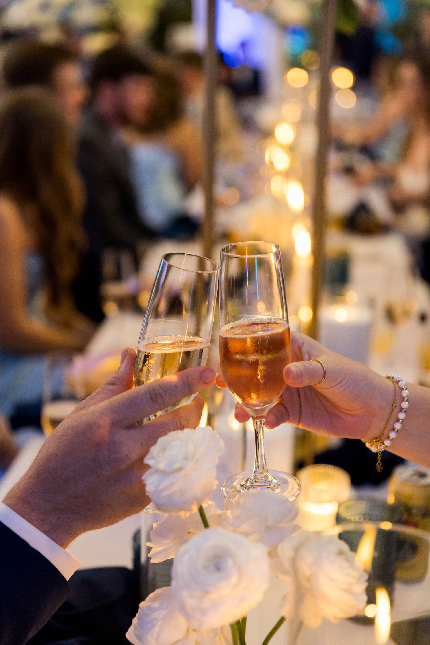Two hands clinking champagne flutes at an elegant evening celebration with soft bokeh lights in the background.