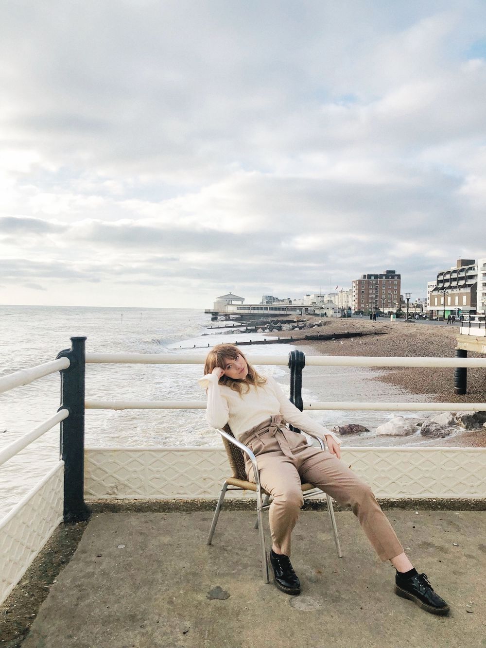 Woman in cream sweater and tan trousers sits on chair by seaside promenade with beach and pier backdrop.