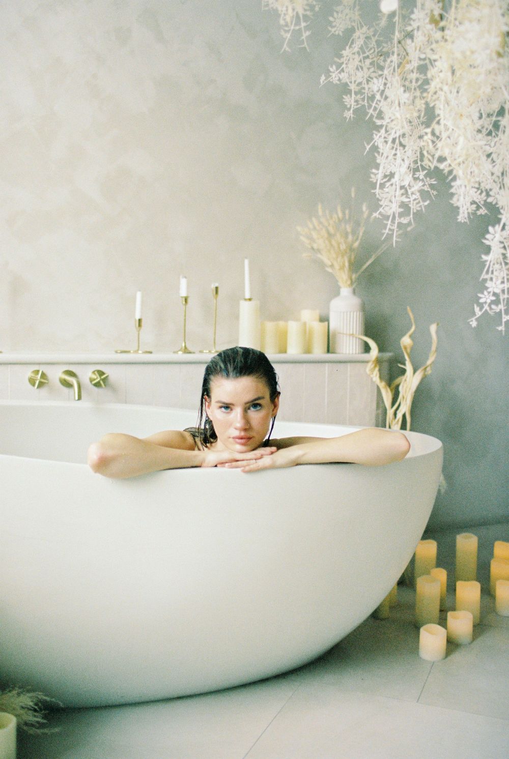 Woman relaxing in a freestanding white bathtub surrounded by candles and dried floral arrangements.