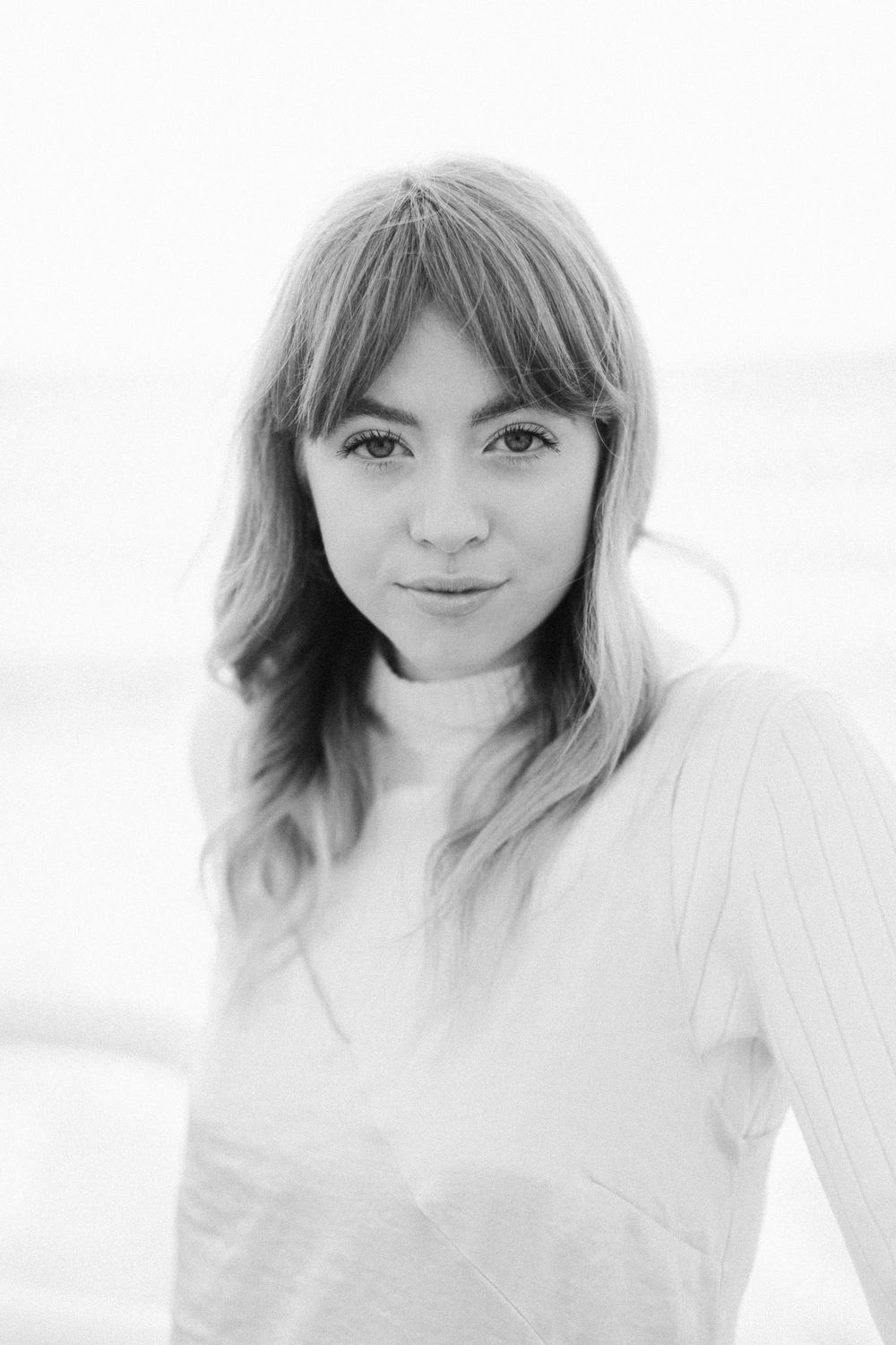 Black and white portrait of a young woman with bangs and wavy hair wearing a light sweater near the ocean.