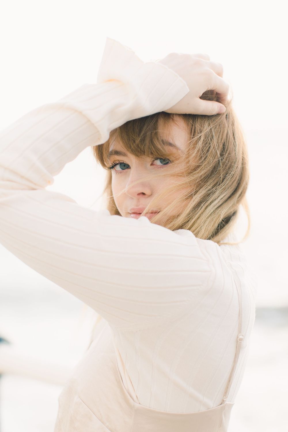 Young woman in cream ribbed top holds hair back, gazing over shoulder in soft natural light.