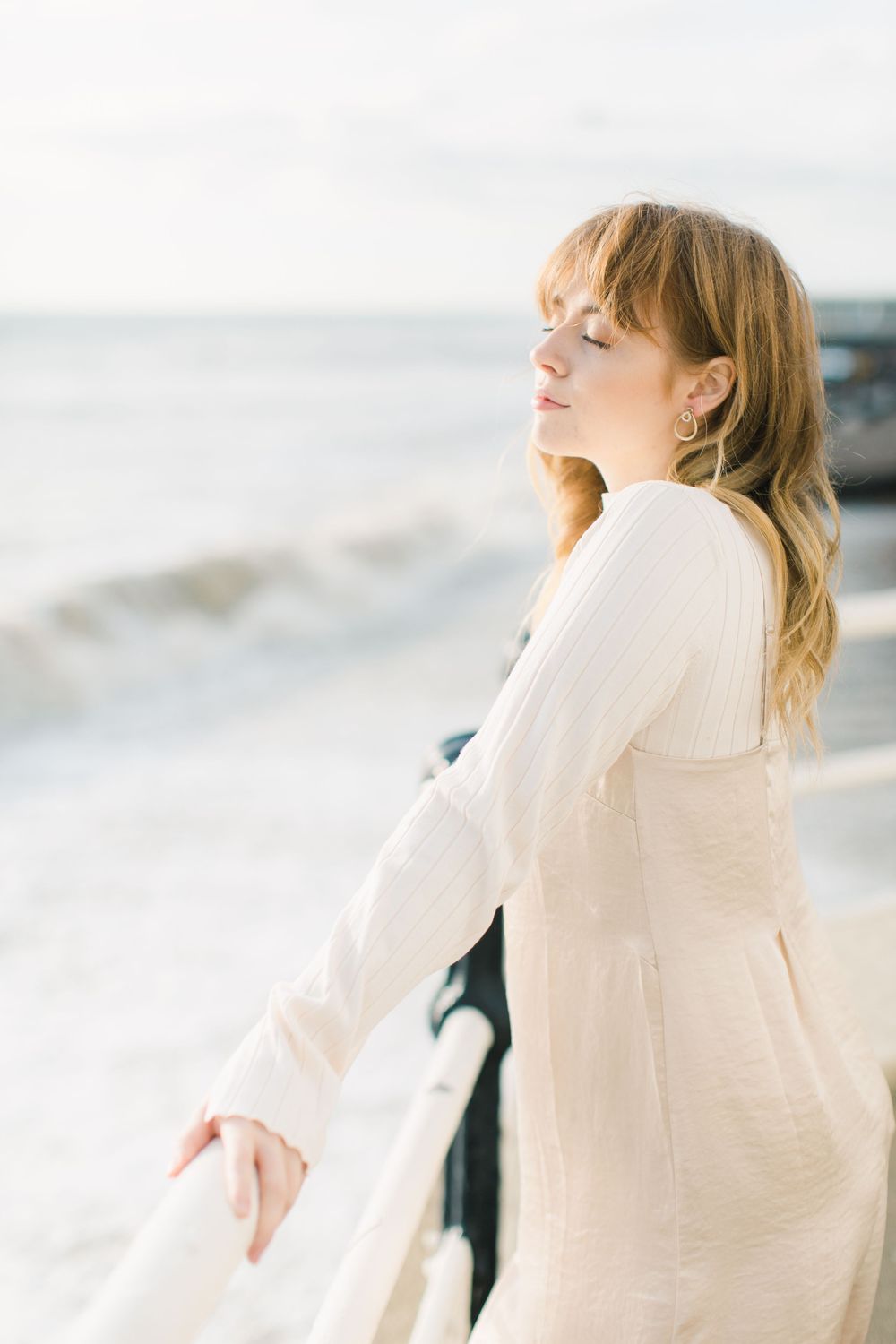 Woman in white linen dress standing by seaside railing with eyes closed, enjoying ocean breeze.