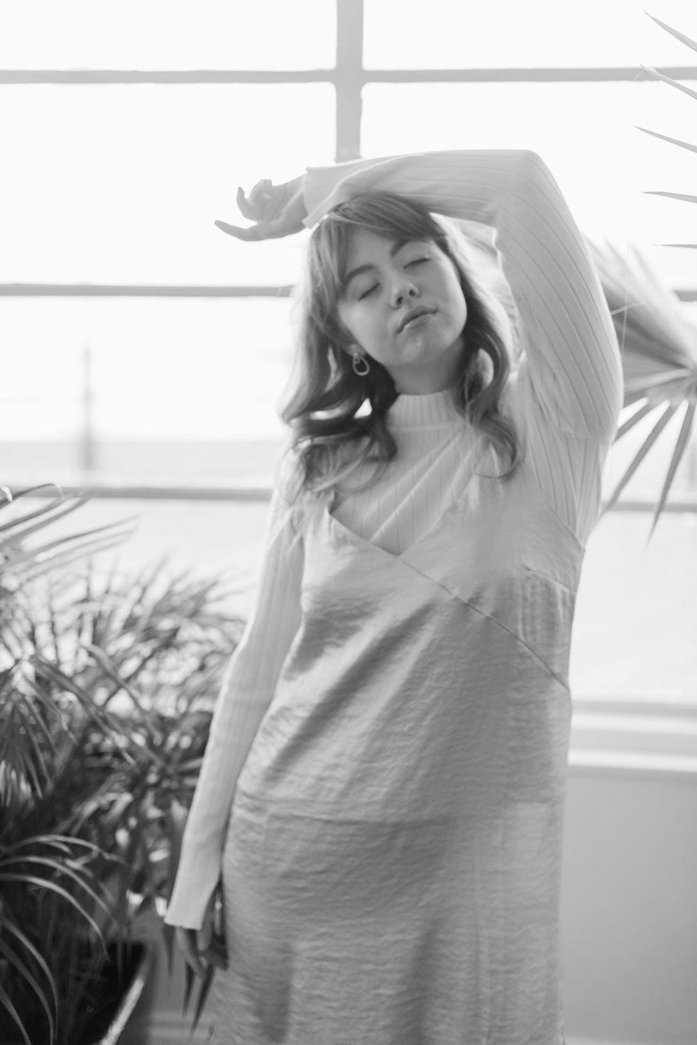 Black and white photo of a woman in a white slip dress posing with arm raised near tropical plants.