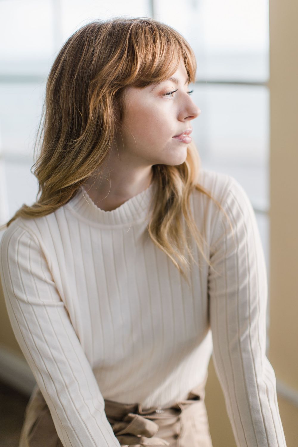 Young woman with bangs in a cream ribbed turtleneck sweater gazing thoughtfully toward a window indoors.