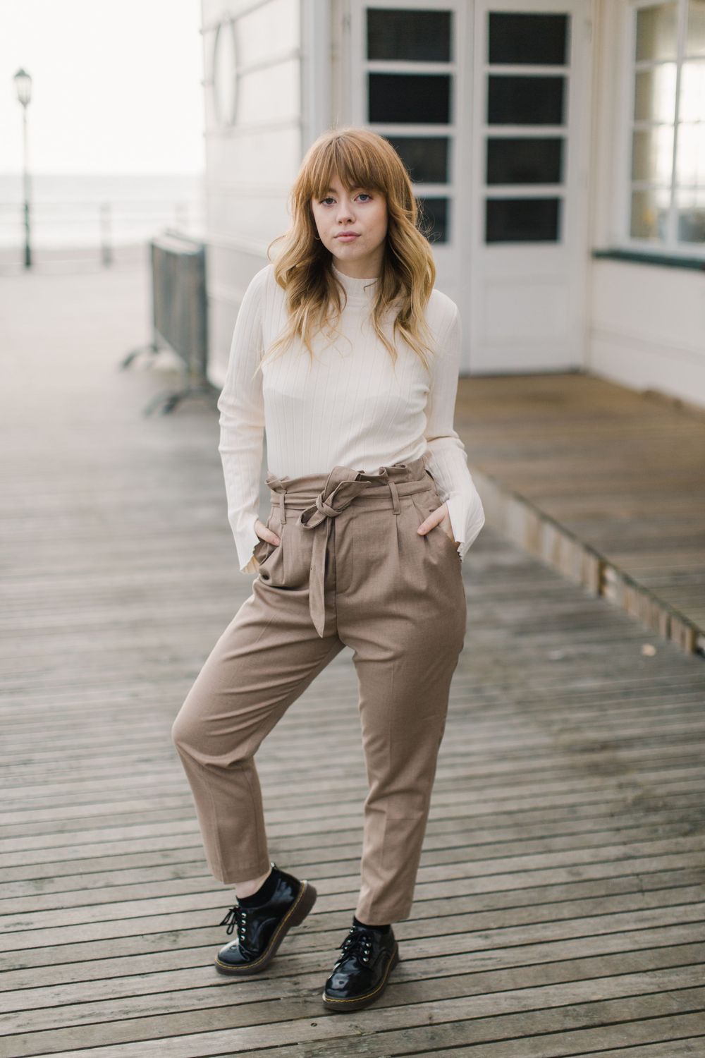 Young woman with bangs wearing a white sweater and tan paperbag trousers with black shoes on a wooden boardwalk.
