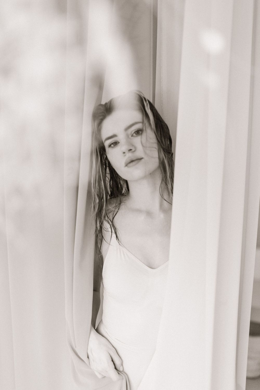 Black and white portrait of a young woman in a white dress gazing through sheer curtains.