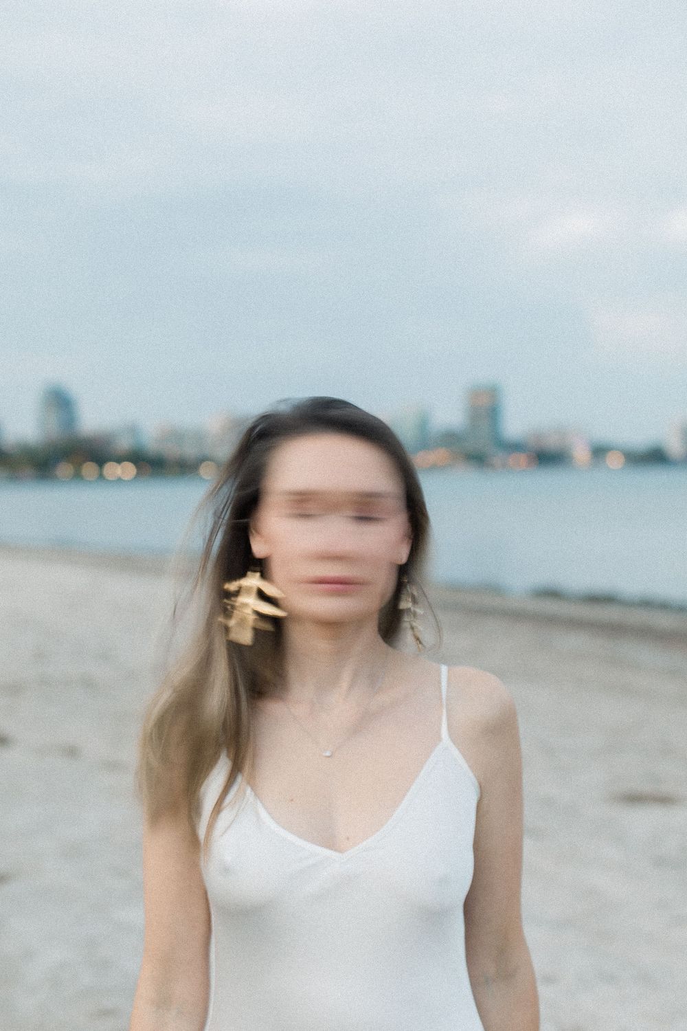 Woman in white dress standing on beach with city skyline in background.