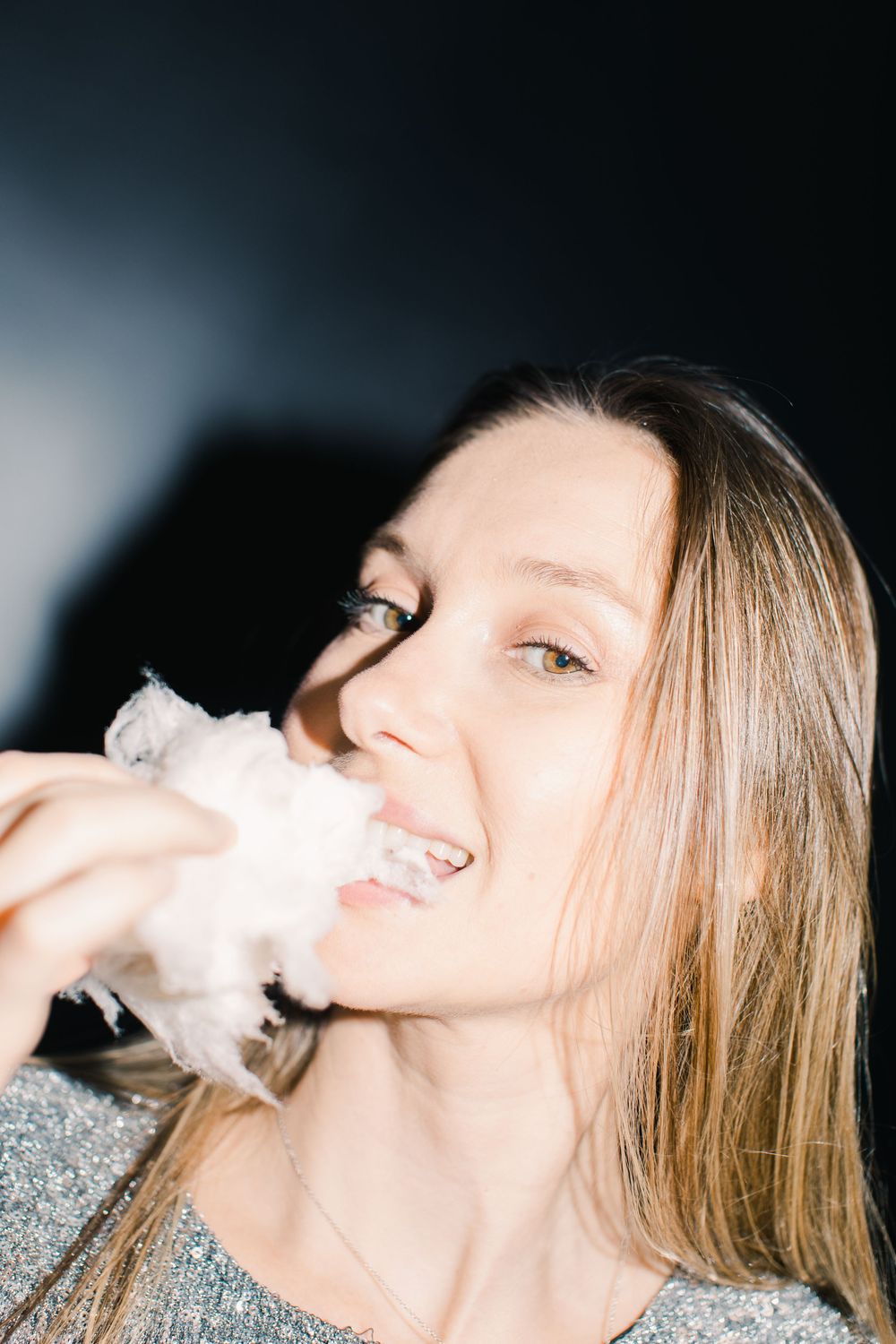 Young woman with long blonde hair smiling while eating white cotton candy against dark background.