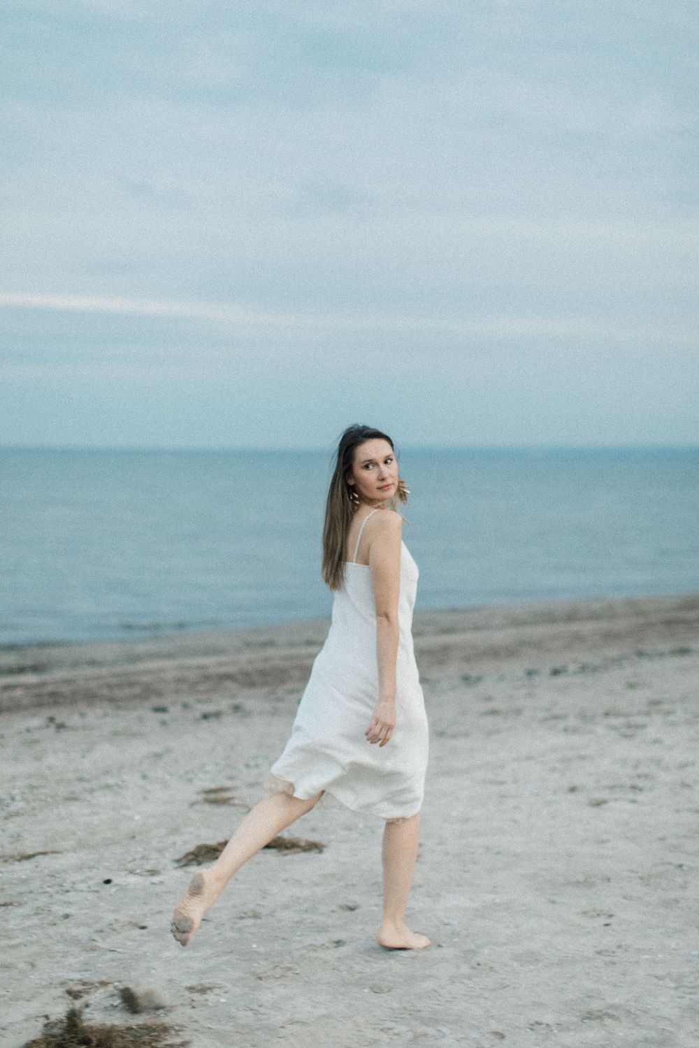 Woman in white sundress walking barefoot on sandy beach with calm ocean and cloudy sky backdrop.