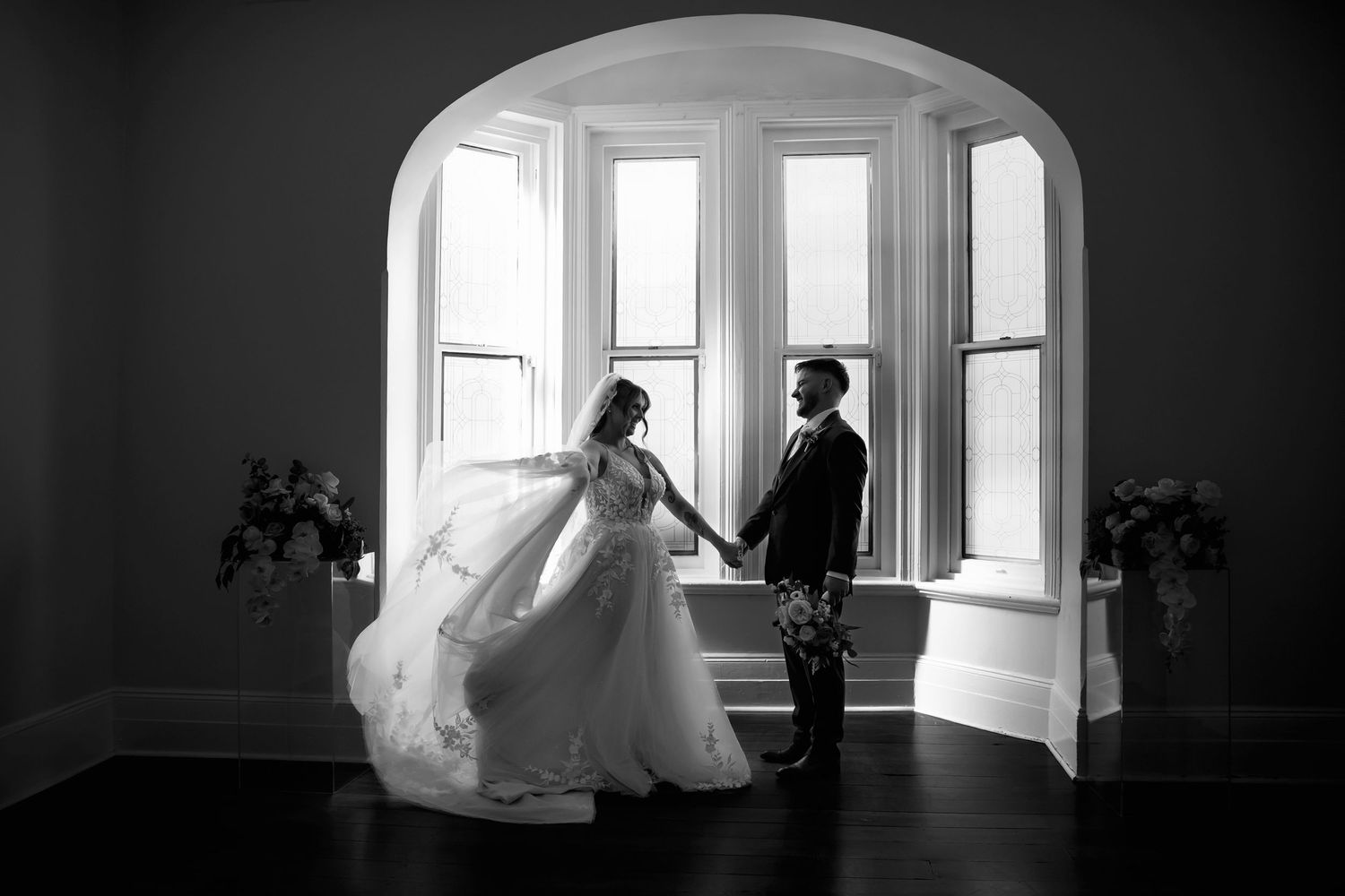 Black and white photo of bride and groom holding hands by arched bay window in elegant venue.