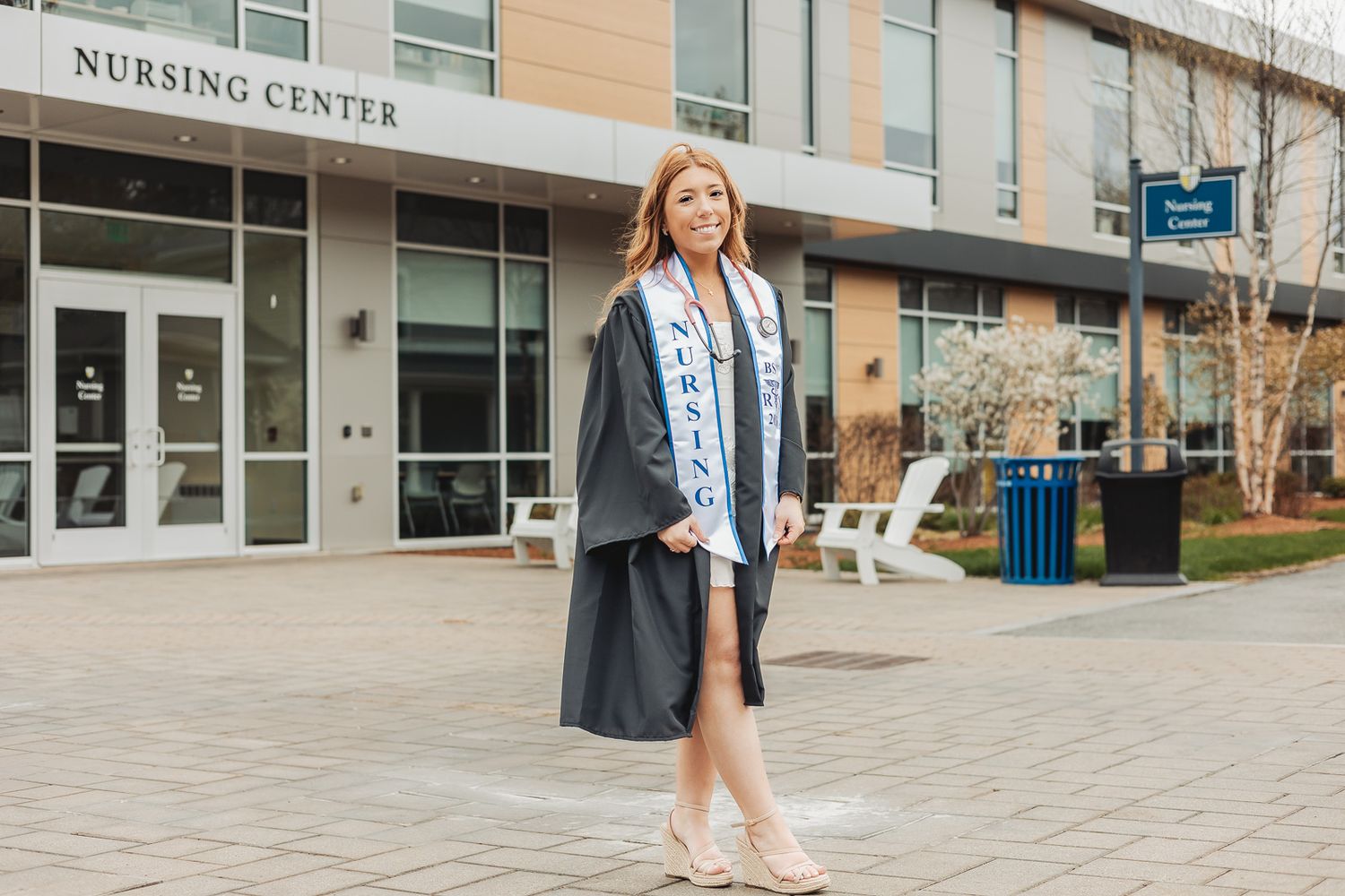 Nursing graduate in cap and gown with stole poses proudly outside Nursing Center building on graduation day.