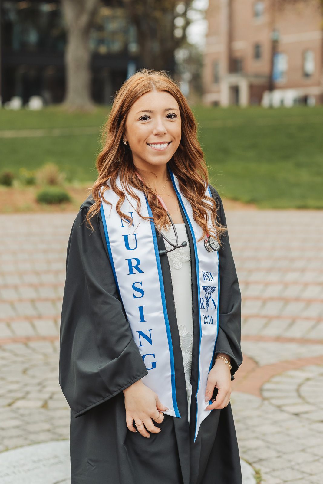 Smiling nursing graduate in black gown and NURSING stole poses outdoors on brick path at college campus.