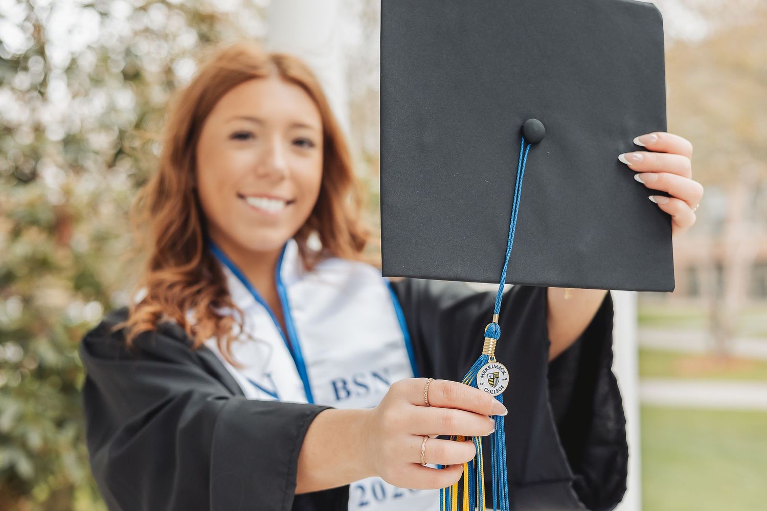 Smiling female graduate in black gown holding up cap with blue tassel and BSN medallion outdoors.